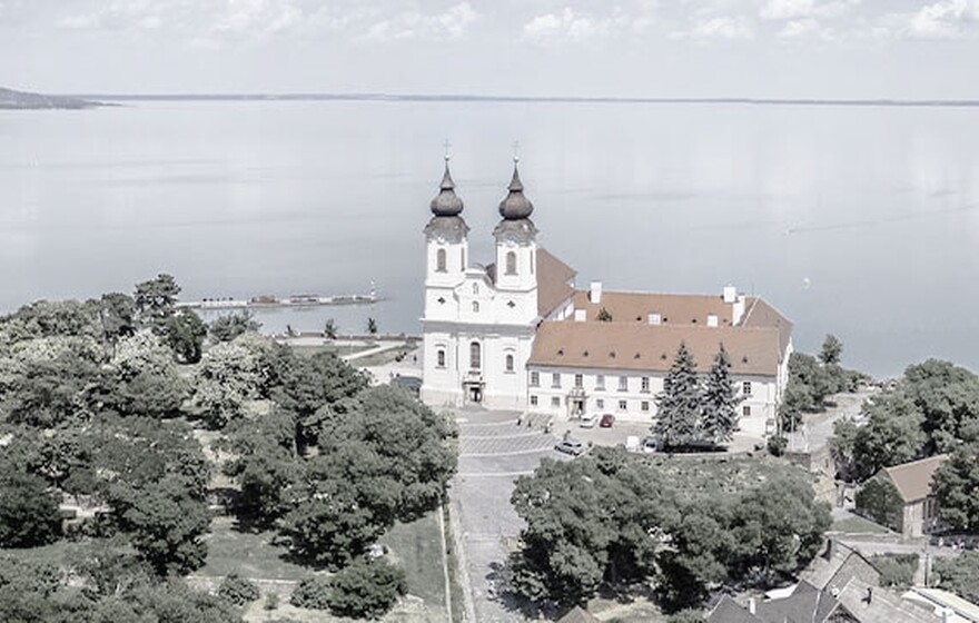 Aerial view of Tihany Abbey with twin towers, red roof, surrounding greenery, and turquoise Lake Balaton in the background.