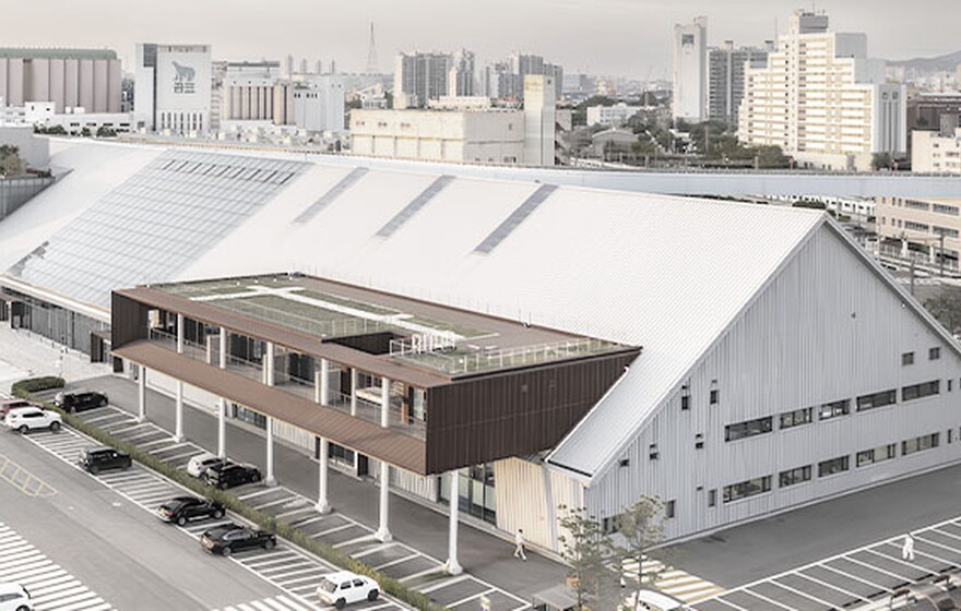 Modern industrial-style building complex with large sloped roofs, rooftop greenery, parking area, and city skyline at sunset.