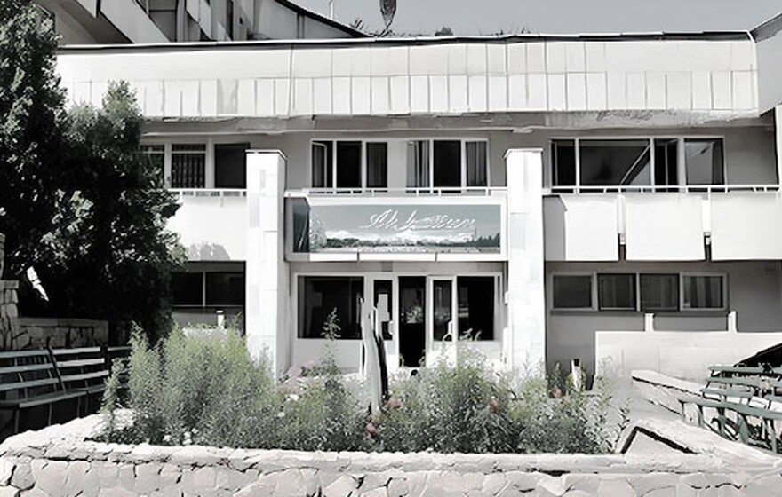 Front view of Ak-Kayyn Sanatorium with stone walls, green benches, and a garden, showing its unique 1970s architecture under clear blue sky.