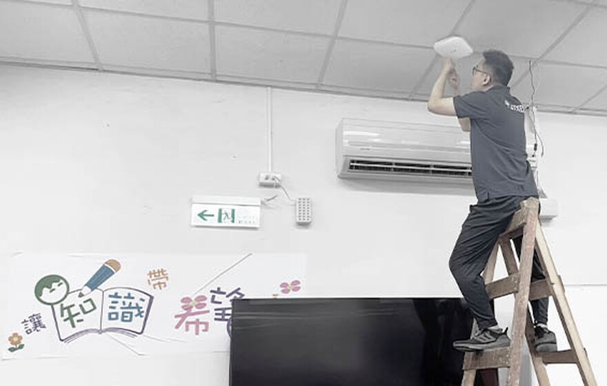 One technician in Zyxel shirts work in a classroom, with one on a ladder installing equipment near the ceiling above a TV and colorful wall poster.