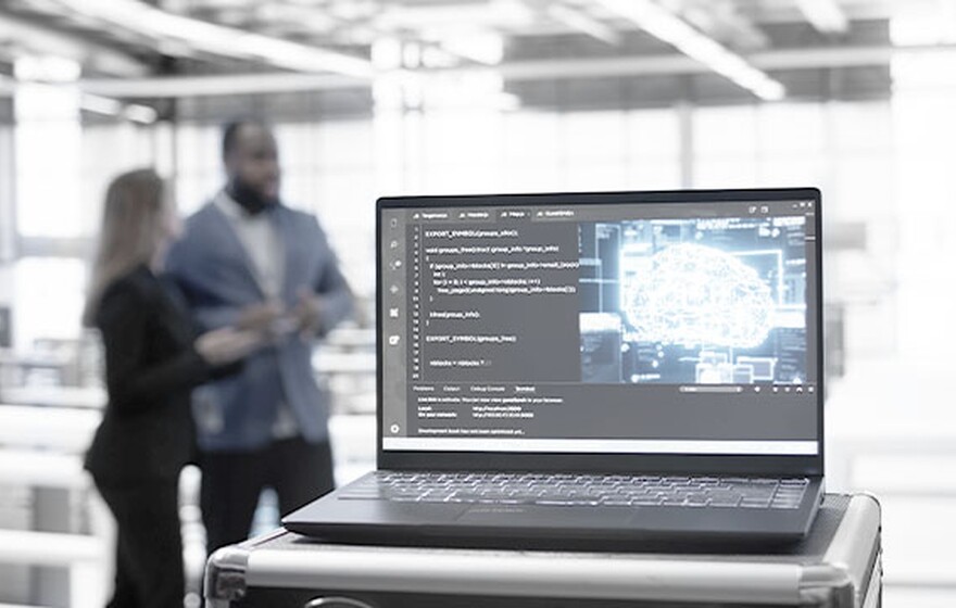 Laptop in a data center displaying code and a glowing digital brain icon with two blurred figures in the background.