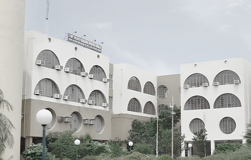 One Indian school building with arched and circular windows, yellow and white facade, lush greenery, and clear blue sky.