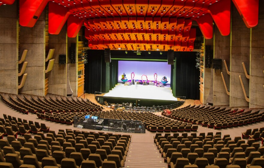 Spacious modern concert hall with red ceiling panels, tiered seating, wooden acoustic design, and a brightly lit stage prepared for performance.