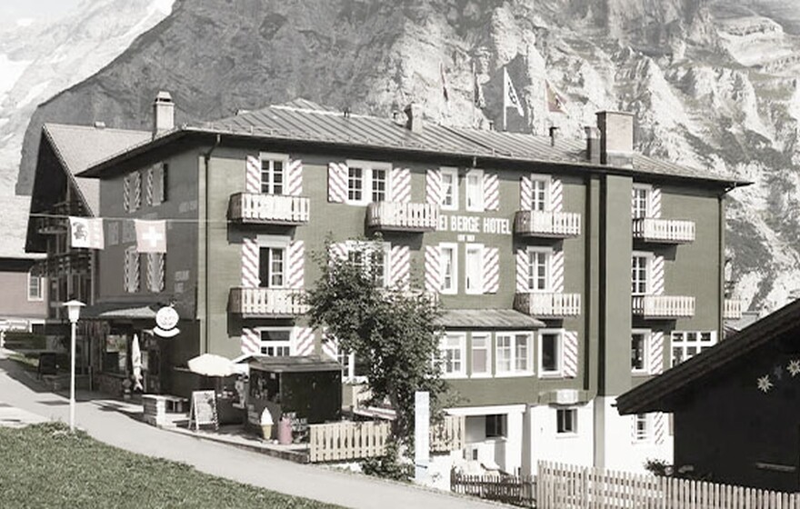 Hotel Drei Berge, a charming green hotel with red-and-white shutters in Mürren village, set against majestic rocky mountains and clear blue sky.