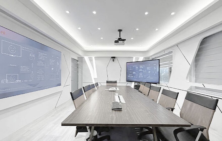 Modern conference room with a long wood table, orange chairs, and dual blue digital screens showing technical AV diagrams.