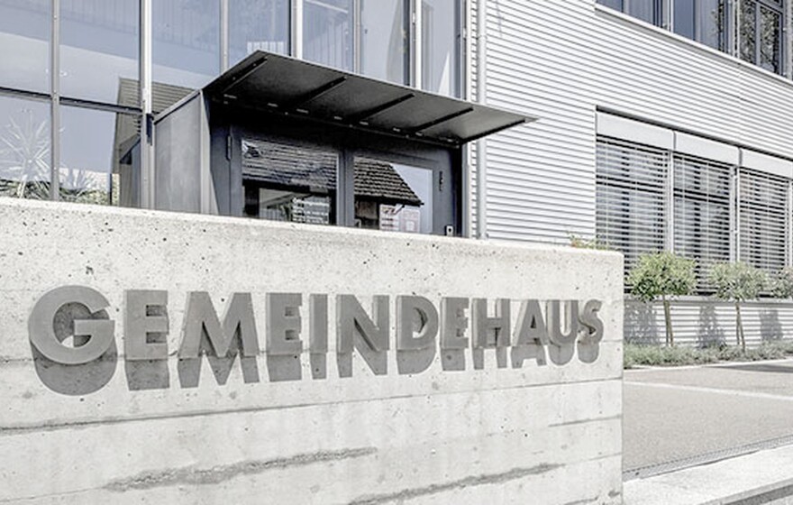 Concrete wall with "GEMEINDEHAUS" lettering in front of a modern industrial building with large windows and small trees.