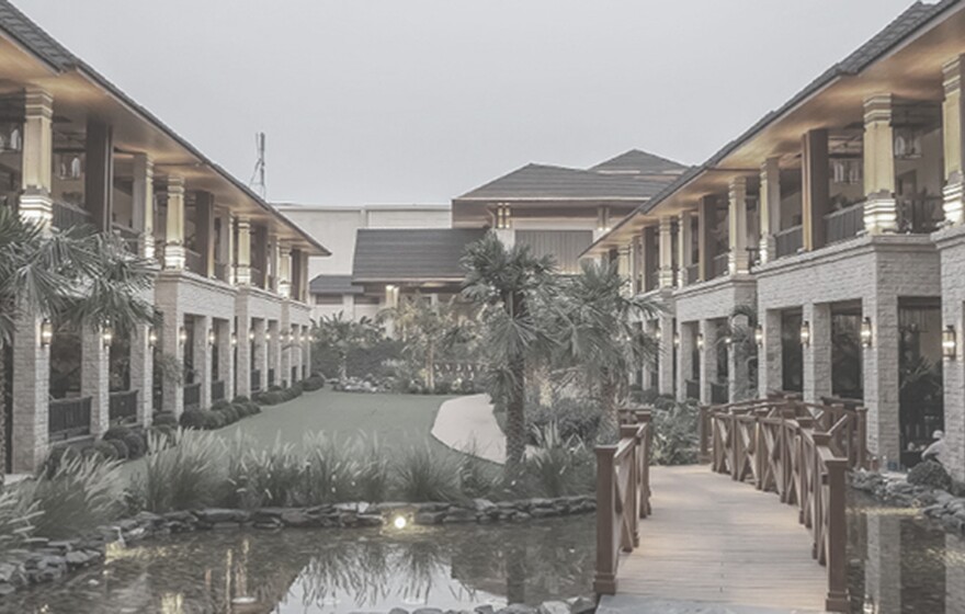 Luxury hotel courtyard with stone facades, warm lighting, balconies, and a wooden bridge over a pond at dusk.