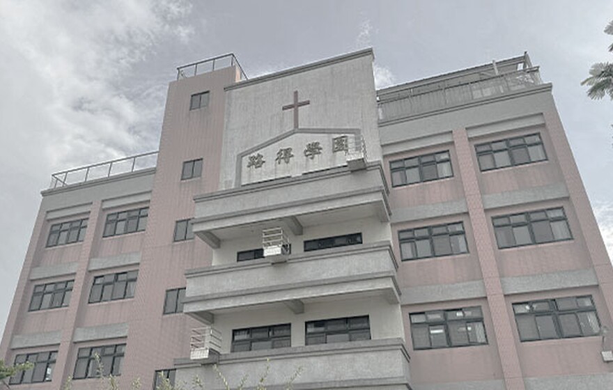 Pink and gray school building with a cross on the central façade, multiple windows, balconies, and a clear sky with light clouds above.