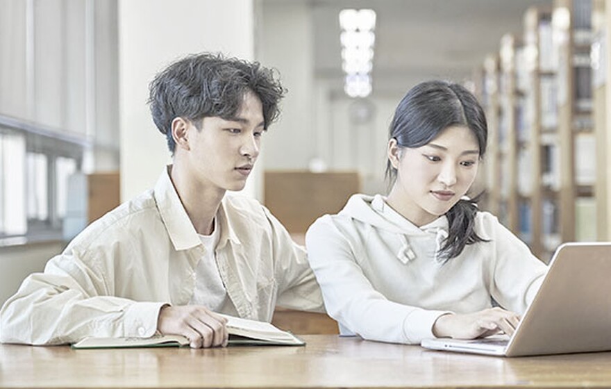 A Korean boy and girl student sit at a wooden library table collaborating with a laptop and an open book.