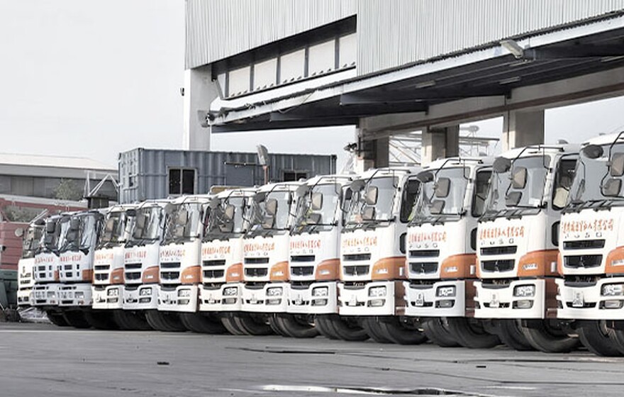 A row of trucks was parked neatly outside a large industrial warehouse under a clear sky, ready for transportation or delivery.