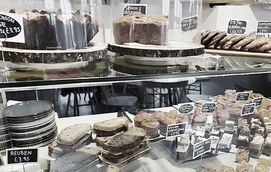 Cafe display with assorted cakes, pastries, brownies, and sandwiches on wooden trays, labeled with handwritten price tags.