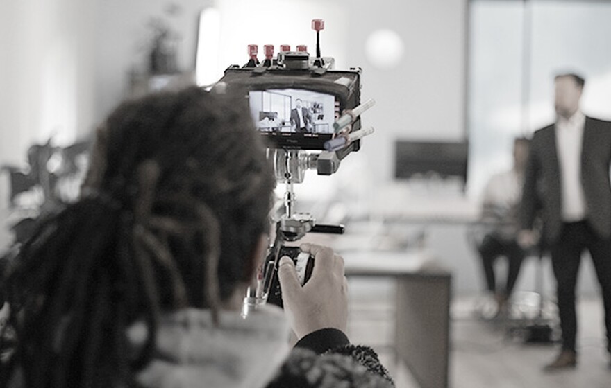 A camera operator films a business scene with actors on set, capturing footage displayed on a monitor during production.