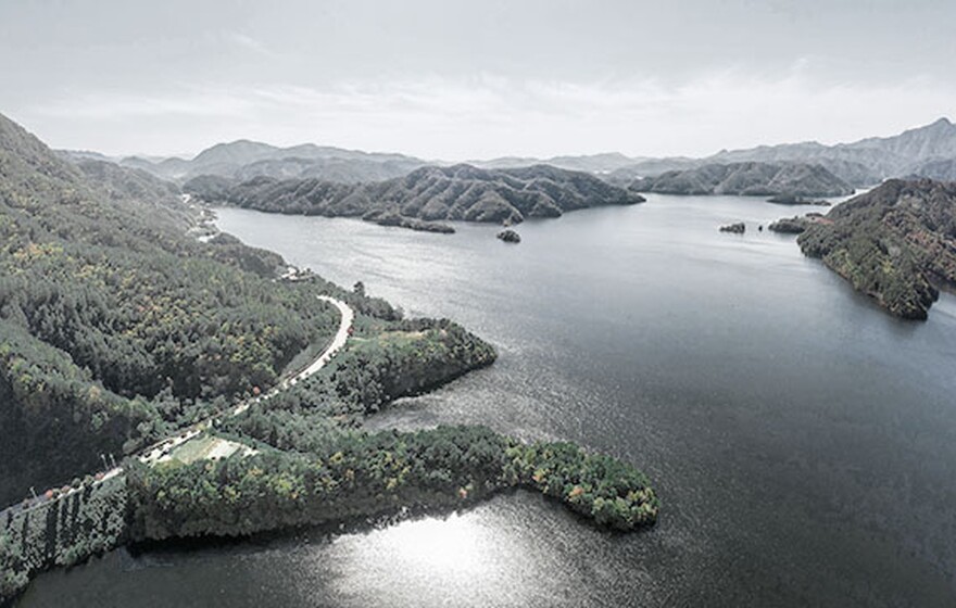 Aerial view of a scenic lake surrounded by forested mountains, winding roads, and clear blue sky on a bright, peaceful day.