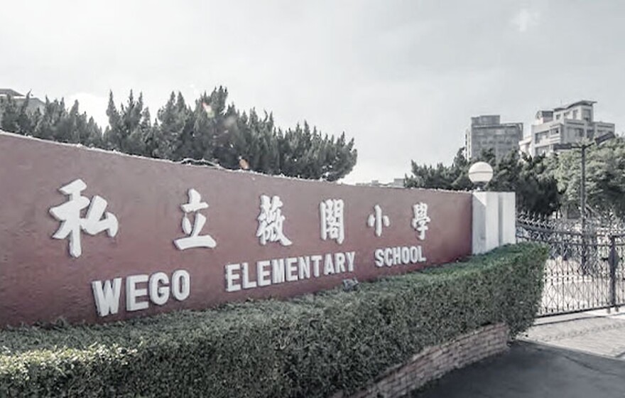 Front entrance of Wego Elementary School with red wall signage in Chinese and English, surrounded by greenery and a gated entry.
