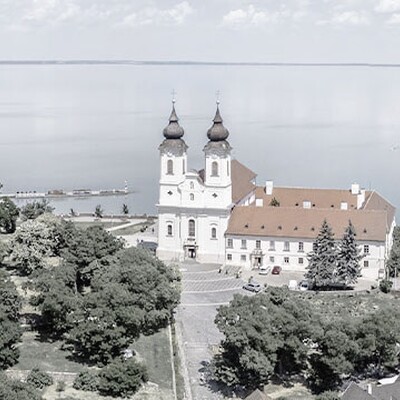 Aerial view of Tihany Abbey with twin towers, red roof, surrounding greenery, and turquoise Lake Balaton in the background.