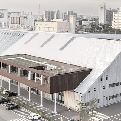 Modern industrial-style building complex with large sloped roofs, rooftop greenery, parking area, and city skyline at sunset.