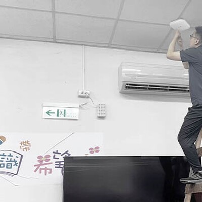 One technician in Zyxel shirts work in a classroom, with one on a ladder installing equipment near the ceiling above a TV and colorful wall poster.
