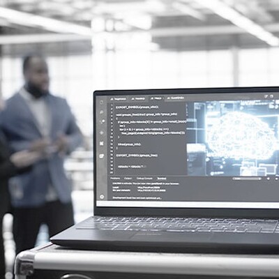 Laptop in a data center displaying code and a glowing digital brain icon with two blurred figures in the background.