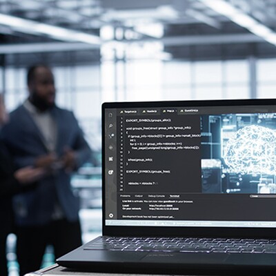 Laptop in a data center displaying code and a glowing digital brain icon with two blurred figures in the background.