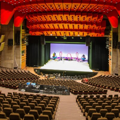 Spacious modern concert hall with red ceiling panels, tiered seating, wooden acoustic design, and a brightly lit stage prepared for performance.
