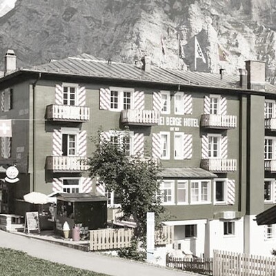 Hotel Drei Berge, a charming green hotel with red-and-white shutters in Mürren village, set against majestic rocky mountains and clear blue sky.