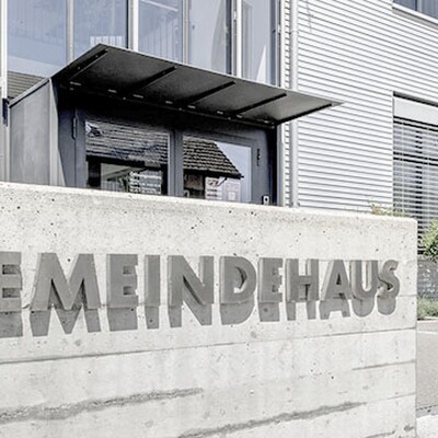 Concrete wall with "GEMEINDEHAUS" lettering in front of a modern industrial building with large windows and small trees.