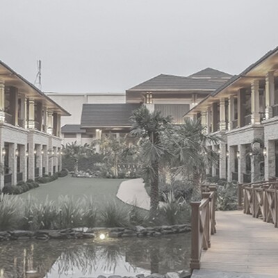 Luxury hotel courtyard with stone facades, warm lighting, balconies, and a wooden bridge over a pond at dusk.