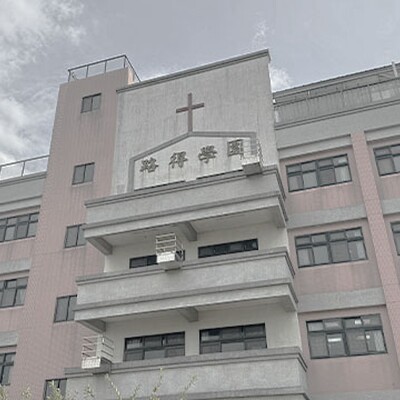 Pink and gray school building with a cross on the central façade, multiple windows, balconies, and a clear sky with light clouds above.