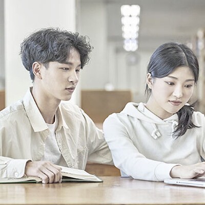 A Korean boy and girl student sit at a wooden library table collaborating with a laptop and an open book.