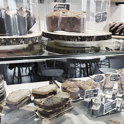 Cafe display with assorted cakes, pastries, brownies, and sandwiches on wooden trays, labeled with handwritten price tags.