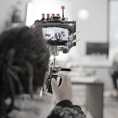 A camera operator films a business scene with actors on set, capturing footage displayed on a monitor during production.