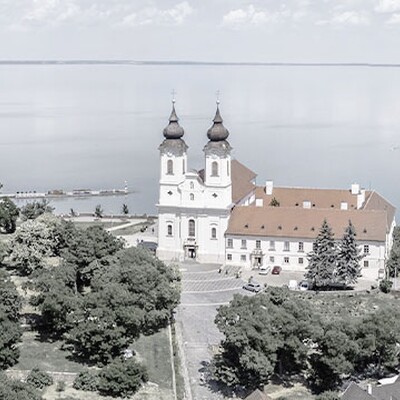 Aerial view of Tihany Abbey with twin towers, red roof, surrounding greenery, and turquoise Lake Balaton in the background.