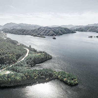 Aerial view of a scenic lake surrounded by forested mountains, winding roads, and clear blue sky on a bright, peaceful day.