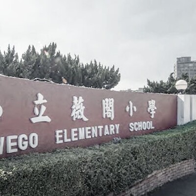 Front entrance of Wego Elementary School with red wall signage in Chinese and English, surrounded by greenery and a gated entry.