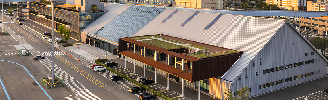 Modern industrial-style building complex with large sloped roofs, rooftop greenery, parking area, and city skyline at sunset.
