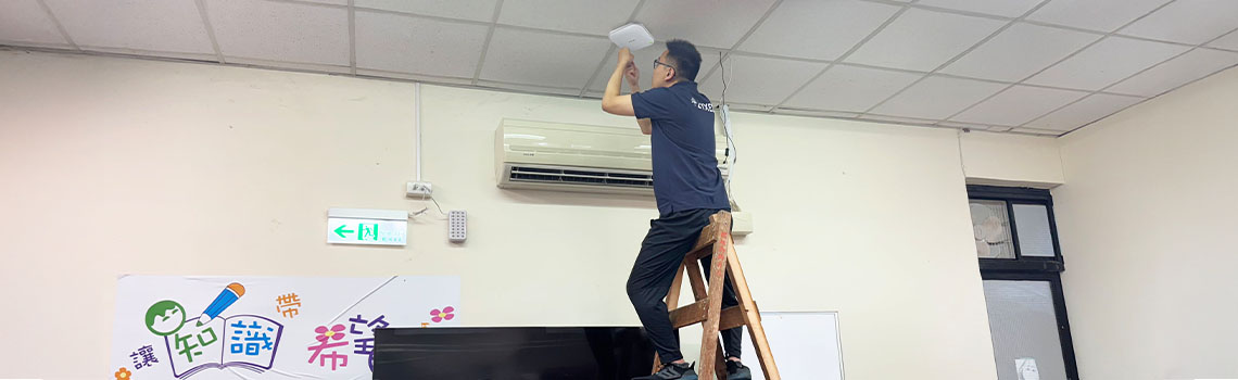 One technician in Zyxel shirts work in a classroom, with one on a ladder installing equipment near the ceiling above a TV and colorful wall poster.
