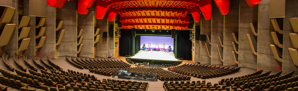 Spacious modern concert hall with red ceiling panels, tiered seating, wooden acoustic design, and a brightly lit stage prepared for performance.

