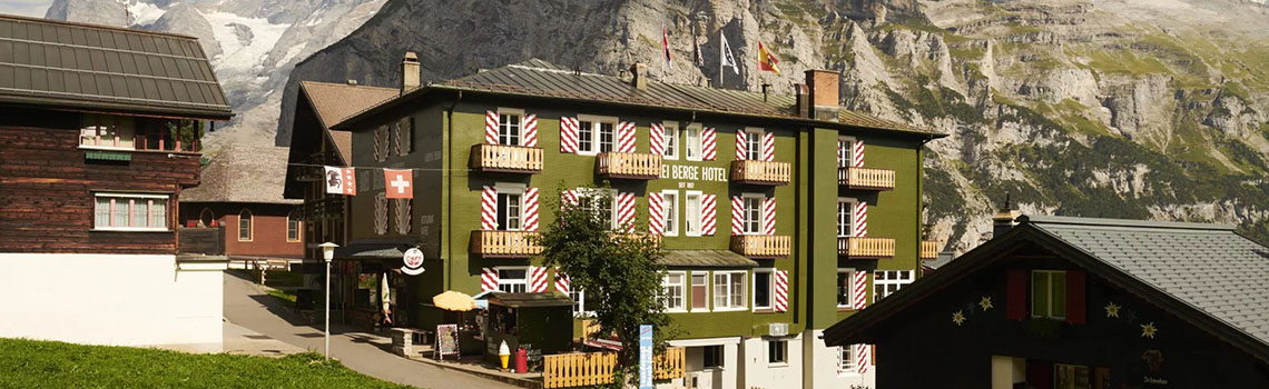 Hotel Drei Berge, a charming green hotel with red-and-white shutters in Mürren village, set against majestic rocky mountains and clear blue sky.