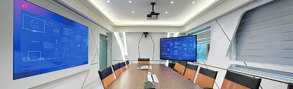 Modern conference room with a long wood table, orange chairs, and dual blue digital screens showing technical AV diagrams.
