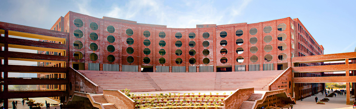 Modern red-brick university building with circular windows, large open amphitheater steps, landscaped courtyard, and students walking around.
