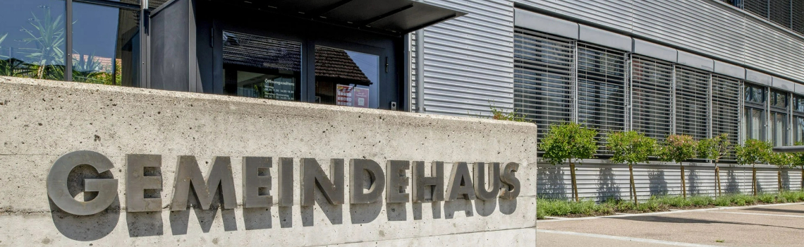 Concrete wall with GEMEINDEHAUS lettering in front of a modern industrial building with large windows and small trees.
