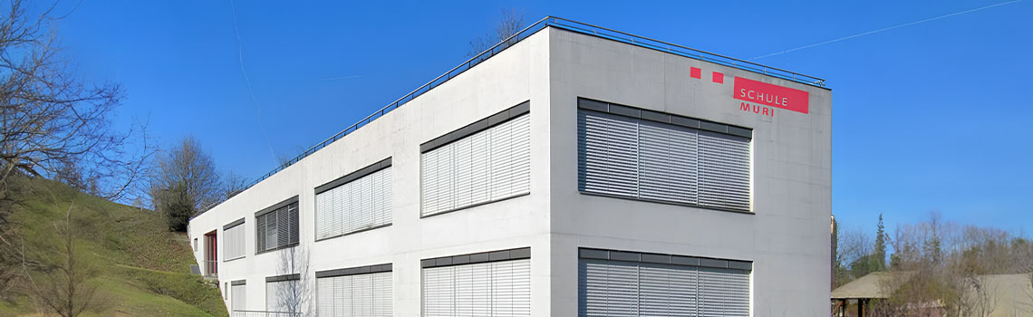 Modern white building of Schule Muri with closed window blinds, set against a clear blue sky and surrounded by grassy slopes.
