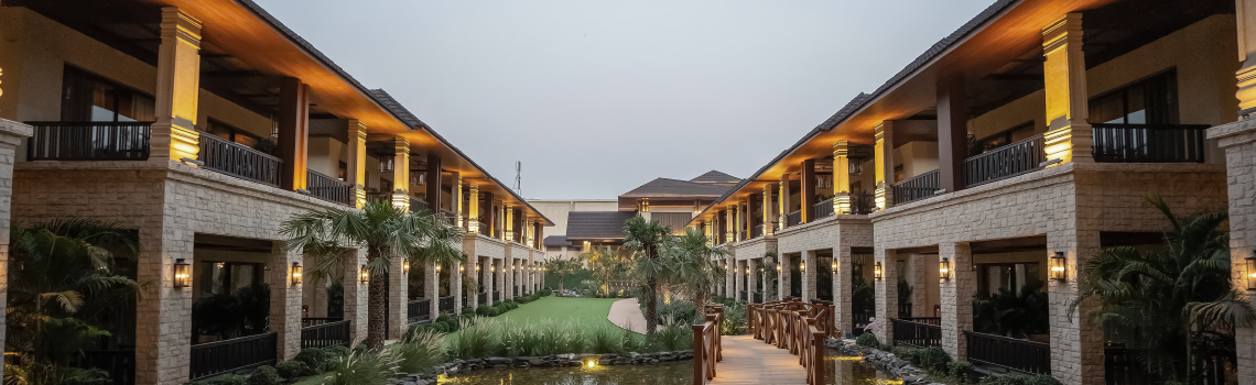 Luxury hotel courtyard with stone facades, warm lighting, balconies, and a wooden bridge over a pond at dusk.