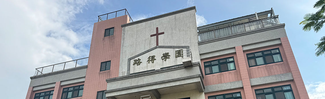 Pink and gray school building with a cross on the central façade, multiple windows, balconies, and a clear sky with light clouds above.
