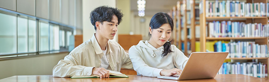 A Korean boy and girl student sit at a wooden library table collaborating with a laptop and an open book.
