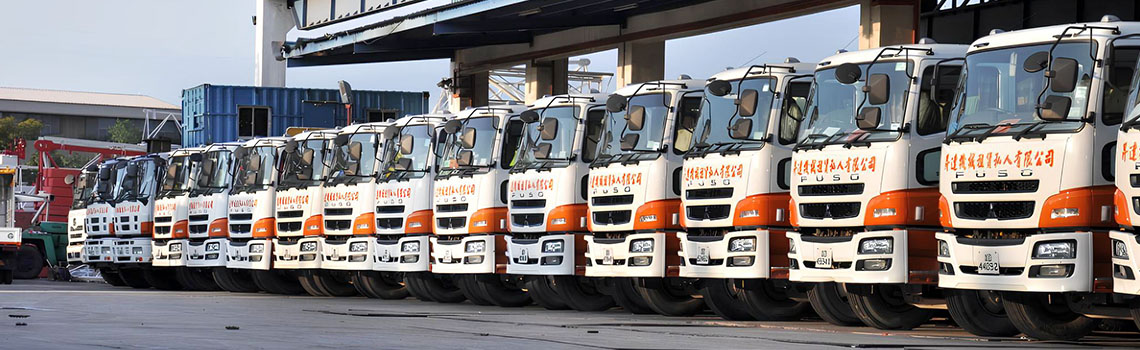A row of trucks was parked neatly outside a large industrial warehouse under a clear sky, ready for transportation or delivery.