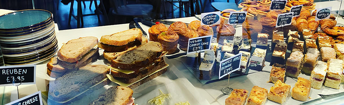 Café display with assorted cakes, pastries, brownies, and sandwiches on wooden trays, labeled with handwritten price tags.