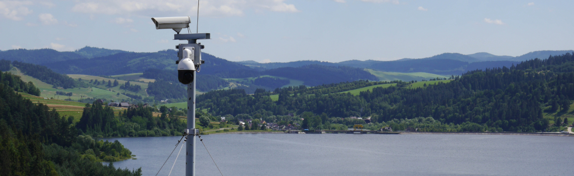 Aerial view of a scenic lake surrounded by forested mountains, winding roads, and clear blue sky on a bright, peaceful day.