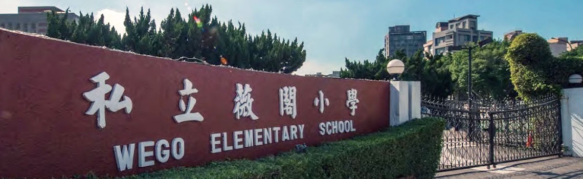 Front entrance of Wego Elementary School with red wall signage in Chinese and English, surrounded by greenery and a gated entry.